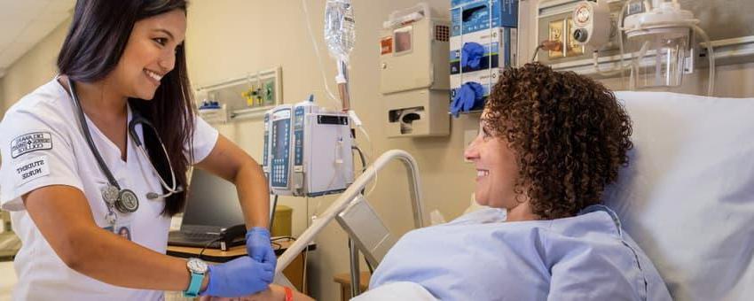 A nursing student in a simulation lab interacts with a patient in a bed.